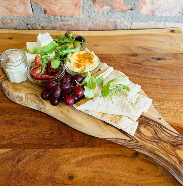 A wooden platter board with a mezze platter containing black and green olives, houmous,  toasted pitta bread, aioli, sundried tomatoes, feta cheese, chilli jam, and grapes arranged on it, placed on a wooden table.